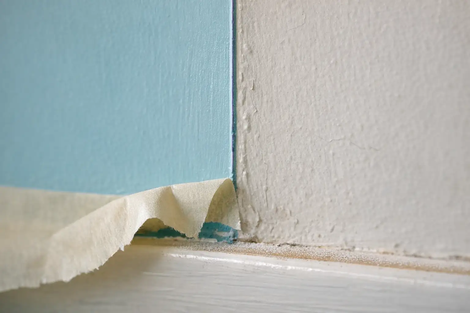 Détail en gros plan d’une coupe de peinture : l’arête parfaitement nette entre un pan fraîchement peint en cyan poudré et un mur blanc cassé, ruban de masquage à moitié décollé, plinthe blanche