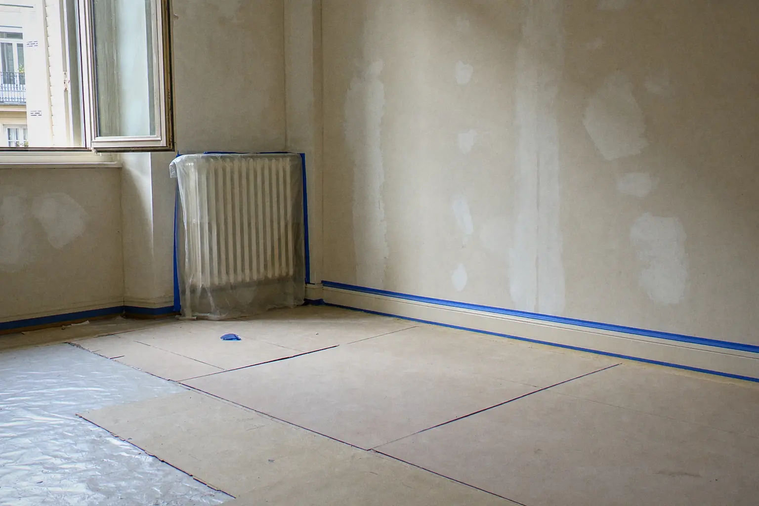 Pièce protégée avant peinture : parquet couvert de bâches et de cartons, radiateur filmé, adhésif bleu le long des plinthes