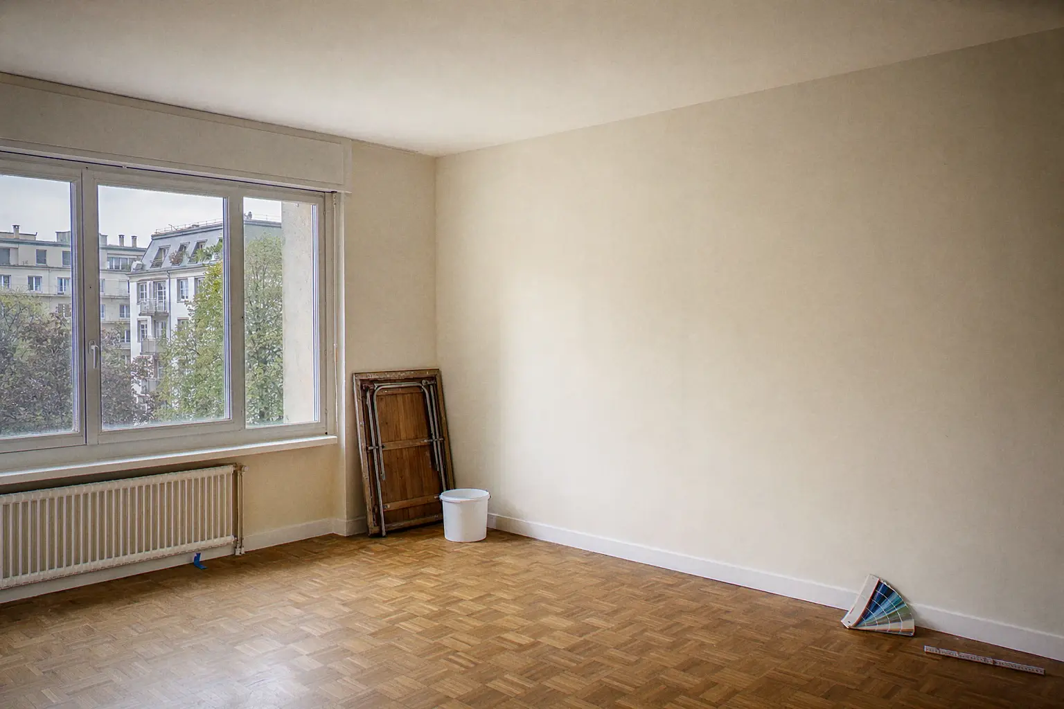 Séjour d'appartement genevois fraîchement repeint et livré : parquet chêne, murs blanc cassé, plafond blanc pur, nuancier et mètre pliant posés contre le mur, table à tapisser pliée au fond