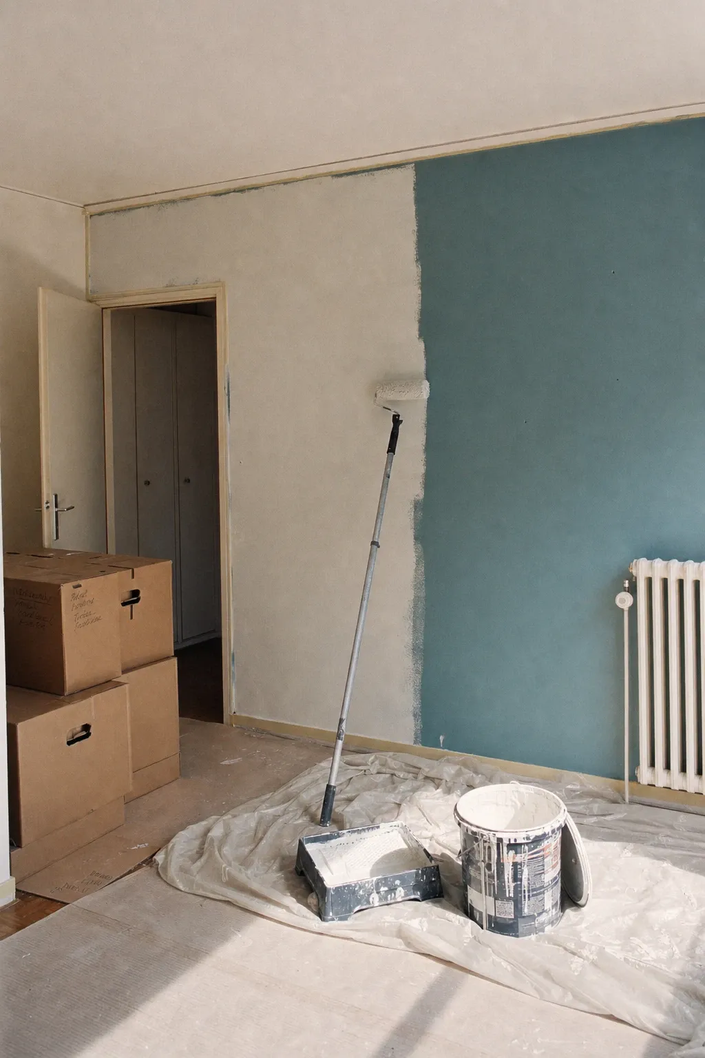 Mur bleu canard à moitié remis en blanc dans un séjour genevois, rouleau sur perche et cartons de déménagement — remise en état avant l’état des lieux de sortie