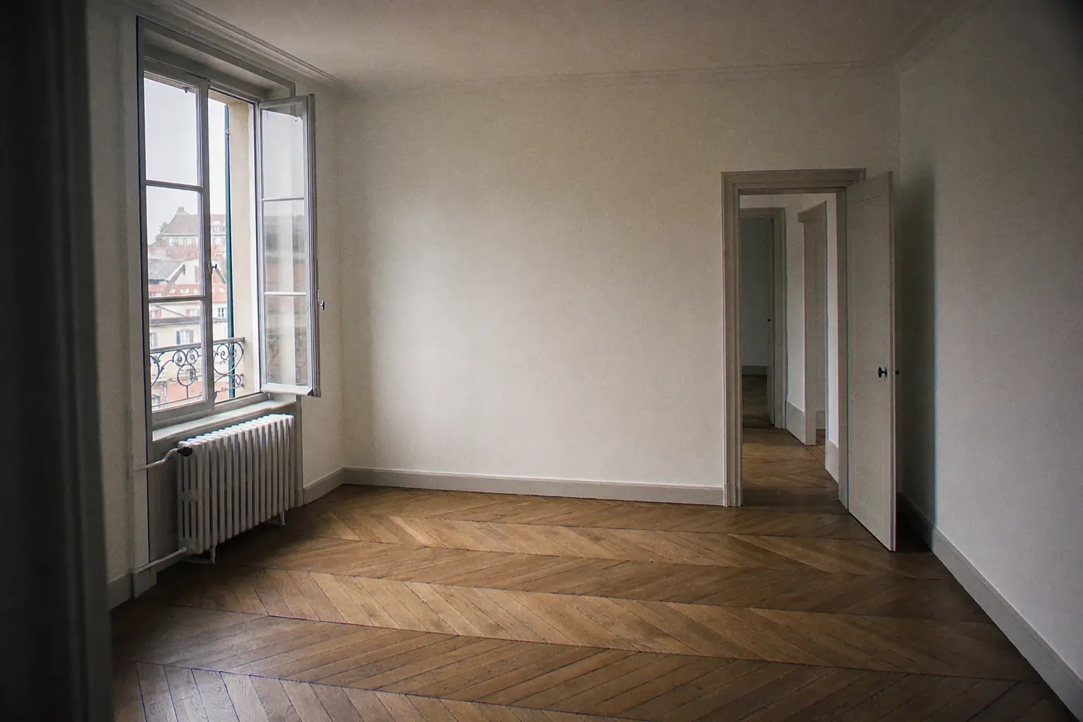 Séjour vide d'appartement ancien remis à neuf entre deux locataires : murs blanc cassé fraîchement repeints, radiateur en fonte repeint, parquet en chevrons, fenêtre entrouverte pour le séchage