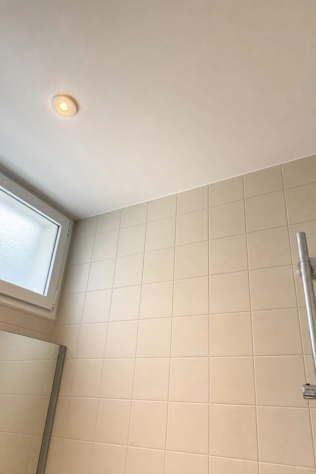 Plafond de salle de bains fraîchement repeint en blanc mat à Genève, jonction nette avec la faïence murale claire, spot encastré et imposte vitrée. Chantier Renovhome SA.